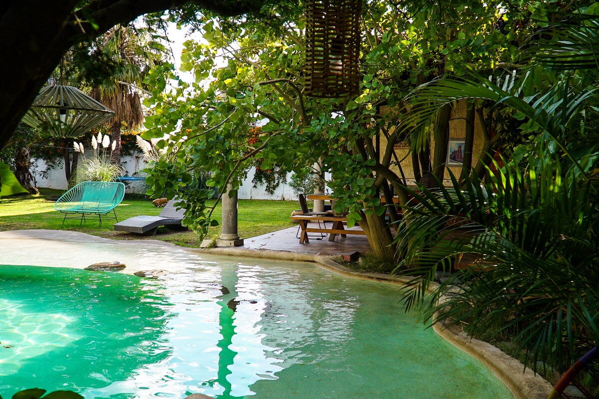 A serene outdoor area features a private pool surrounded by tropical greenery. A shaded seating area and loungers are visible, inviting relaxation. The pool's gentle curve is complemented by a rustic gazebo, creating a harmonious blend of nature and comfort.