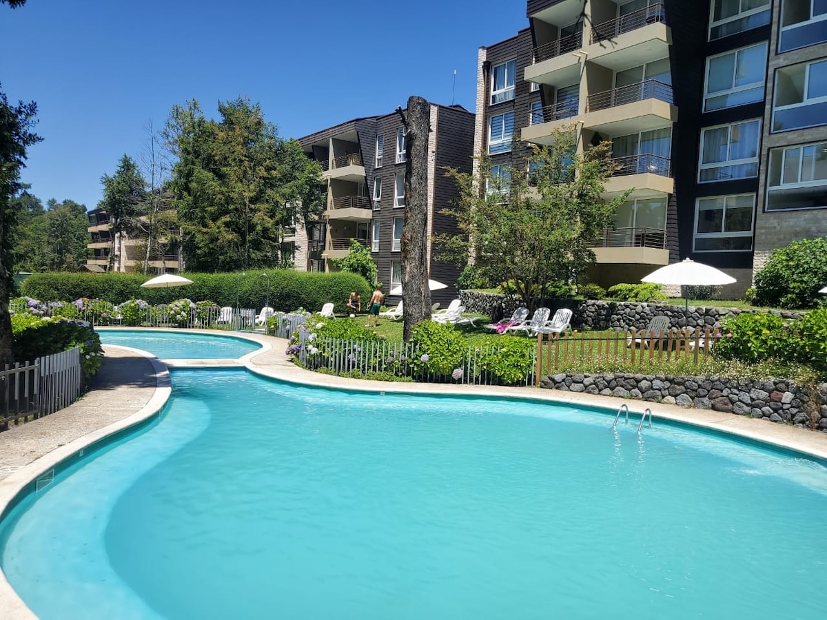 A scenic view of a meandering swimming pool is framed by lush greenery and lounge chairs. The clear blue water reflects the sunlight, while the surrounding buildings feature large windows and balconies, blending harmoniously with the tranquil outdoor space.