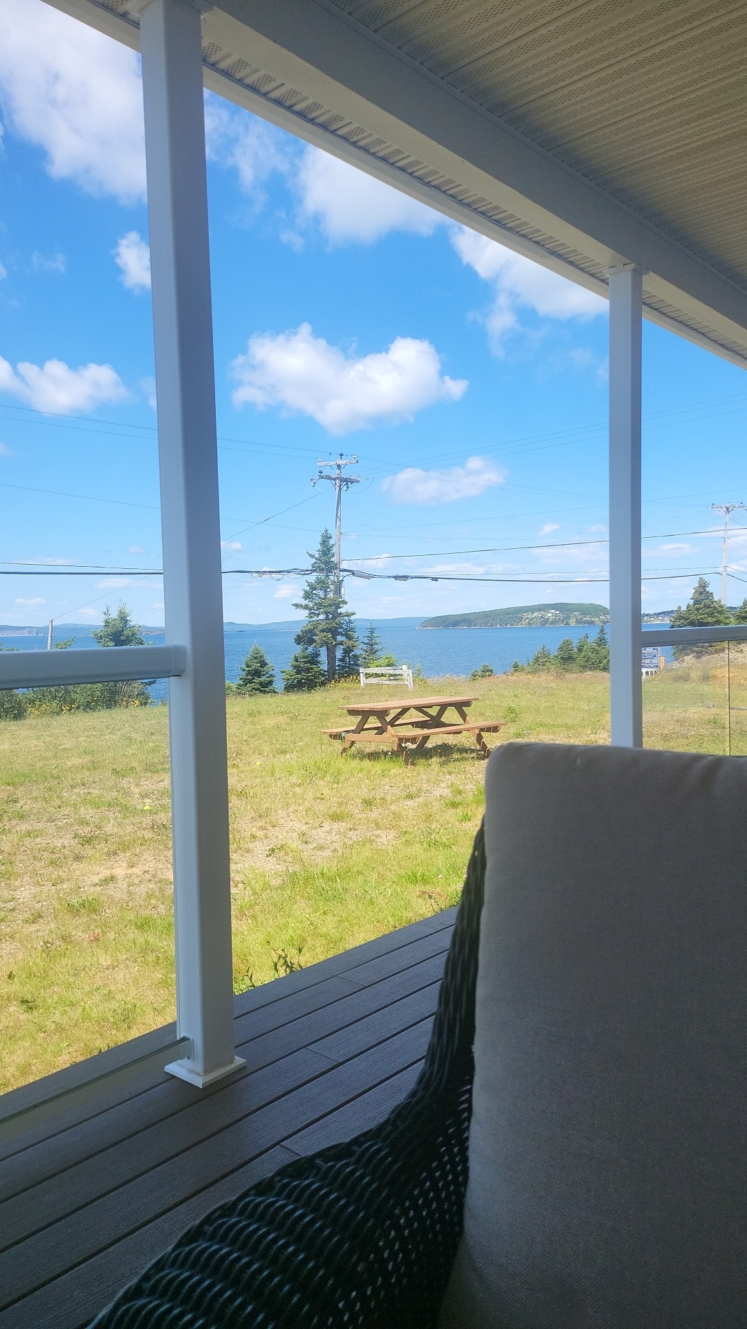 A view of the bay is captured from the large front porch, framed by white railings. Picnic tables are visible on the grassy area, with trees lining the background under a bright blue sky adorned with fluffy clouds.