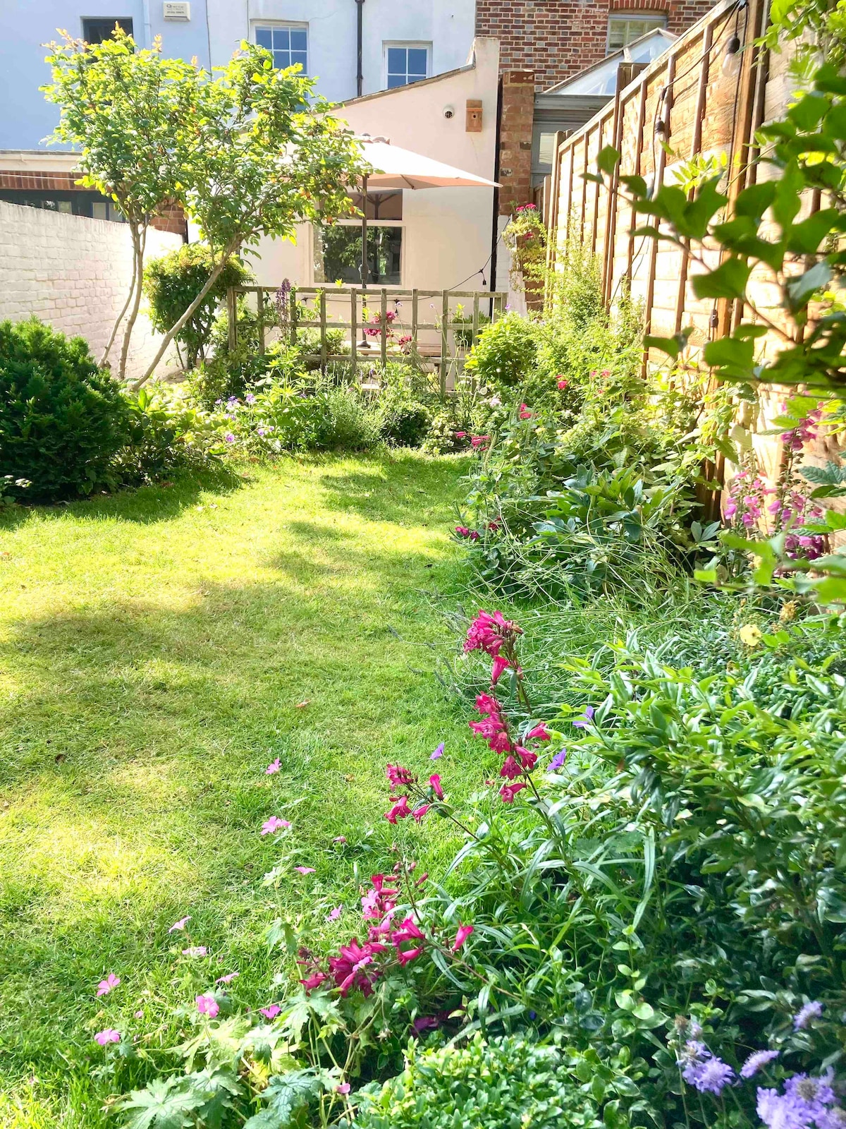 A tranquil garden space is showcased, featuring lush green grass and vibrant floral arrangements. Several plants and small trees frame the area, offering a natural ambiance. In the background, a wooden gate leads to a cozy seating area, adding to the garden's charm.
