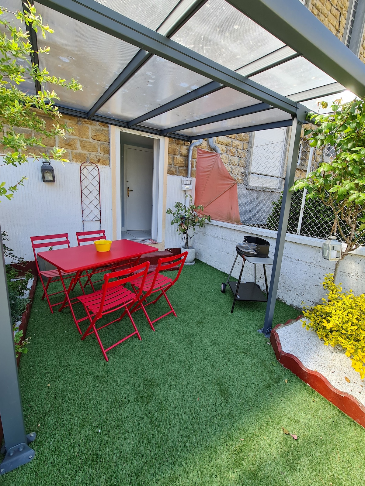An outdoor seating area features a red table surrounded by matching foldable chairs. A small grill is positioned nearby on the artificial grass. Lush greenery and yellow plants frame the space, creating a vibrant and inviting environment.