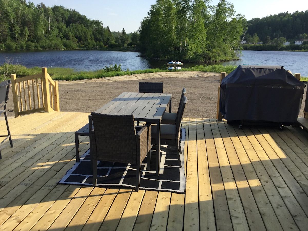 An outdoor deck is shown with a dining table surrounded by six chairs. A grill is positioned nearby among greenery, with a view of the water in the background. The area features a light wood floor and a black and white rug.