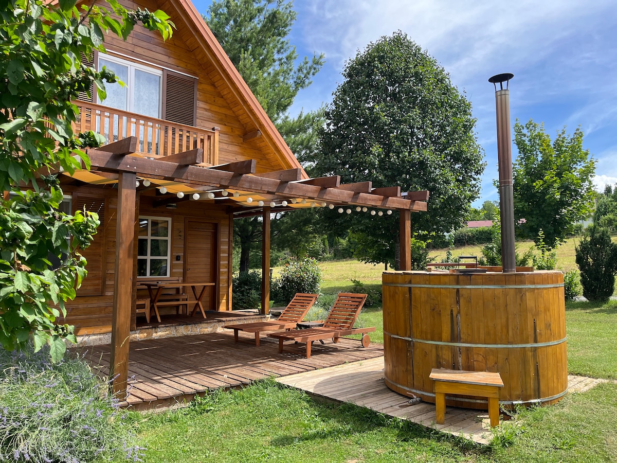 A wooden house features a covered deck with lounge chairs, surrounded by greenery. A wooden hot tub is positioned on the patio, complemented by a seating area. The landscape includes trees and an open sky, adding to the serene atmosphere of the estate.