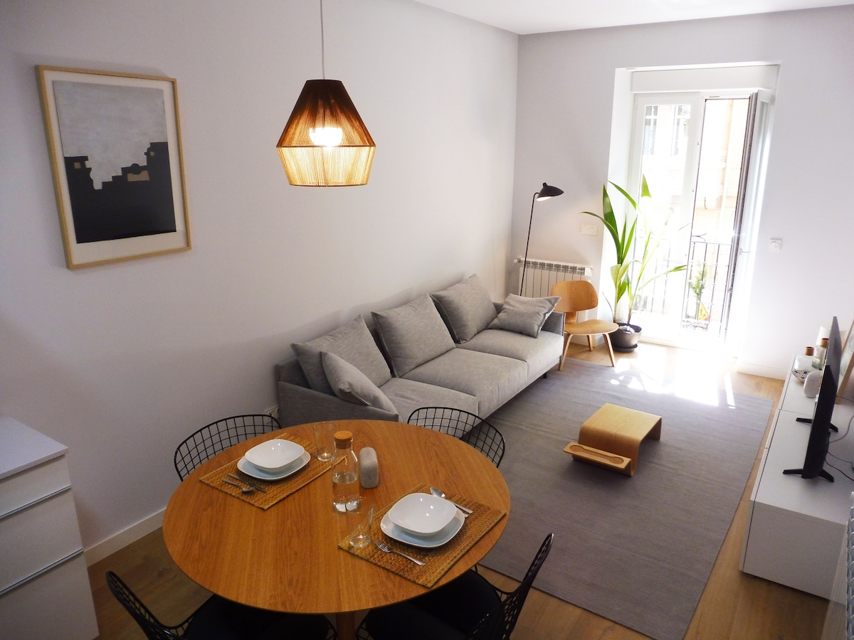 A modern living space is presented with a round wooden dining table set for two and surrounded by black wire chairs. A cozy gray sofa is positioned near a window, complemented by a contemporary floor lamp and a decorative green plant, enhancing the room's inviting atmosphere.