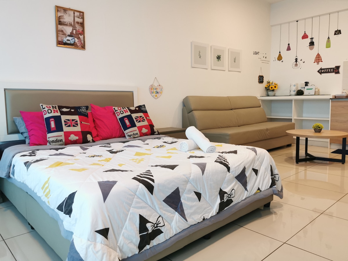 A spacious studio room features a large bed adorned with decorative pillows. A folded couch is positioned nearby, complemented by a small coffee table. Wall decor includes framed art and hanging lights, enhancing the room's cozy atmosphere.