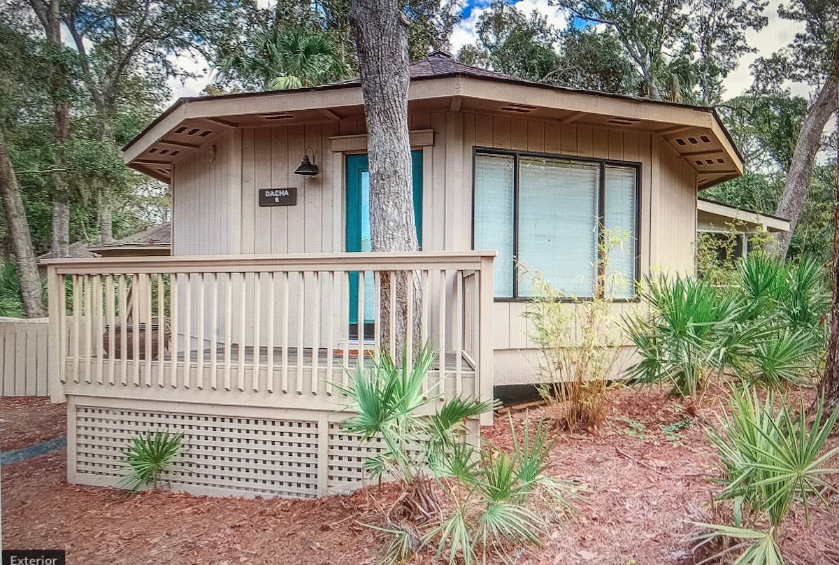 The exterior of the villa features a modern design with natural wood siding. A spacious deck is adorned with a railing and small plants, providing a welcoming entrance. Large windows allow natural light into the home, surrounded by lush greenery.
