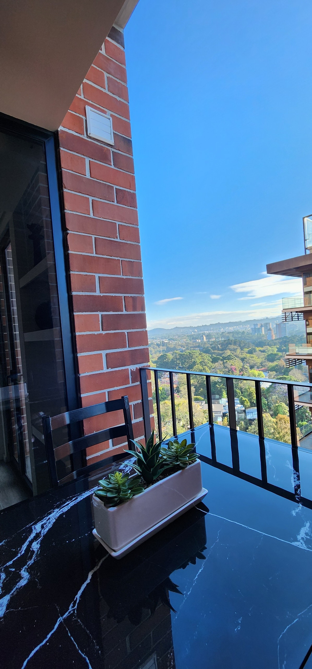 A balcony view features a sleek black marble table adorned with a potted plant. The backdrop presents a vast landscape with distant hills under a clear blue sky, and a glimpse of the city skyline is visible in the distance.