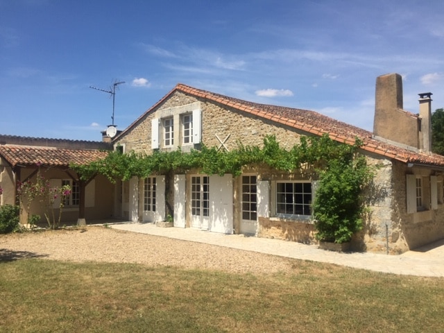 A charming stone house is framed by greenery and displays large windows with shutters. A well-maintained yard features a gravel pathway leading to the entrance. The sky is clear and sunny, enhancing the welcoming appearance of the home.