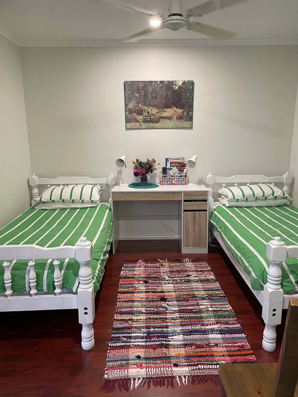 A cozy room features two single beds with green-striped bedding, positioned on polished wooden flooring. A small desk with storage is placed between the beds. A colorful, woven rug adds texture, while a nature-themed artwork hangs above the desk, complementing the space.