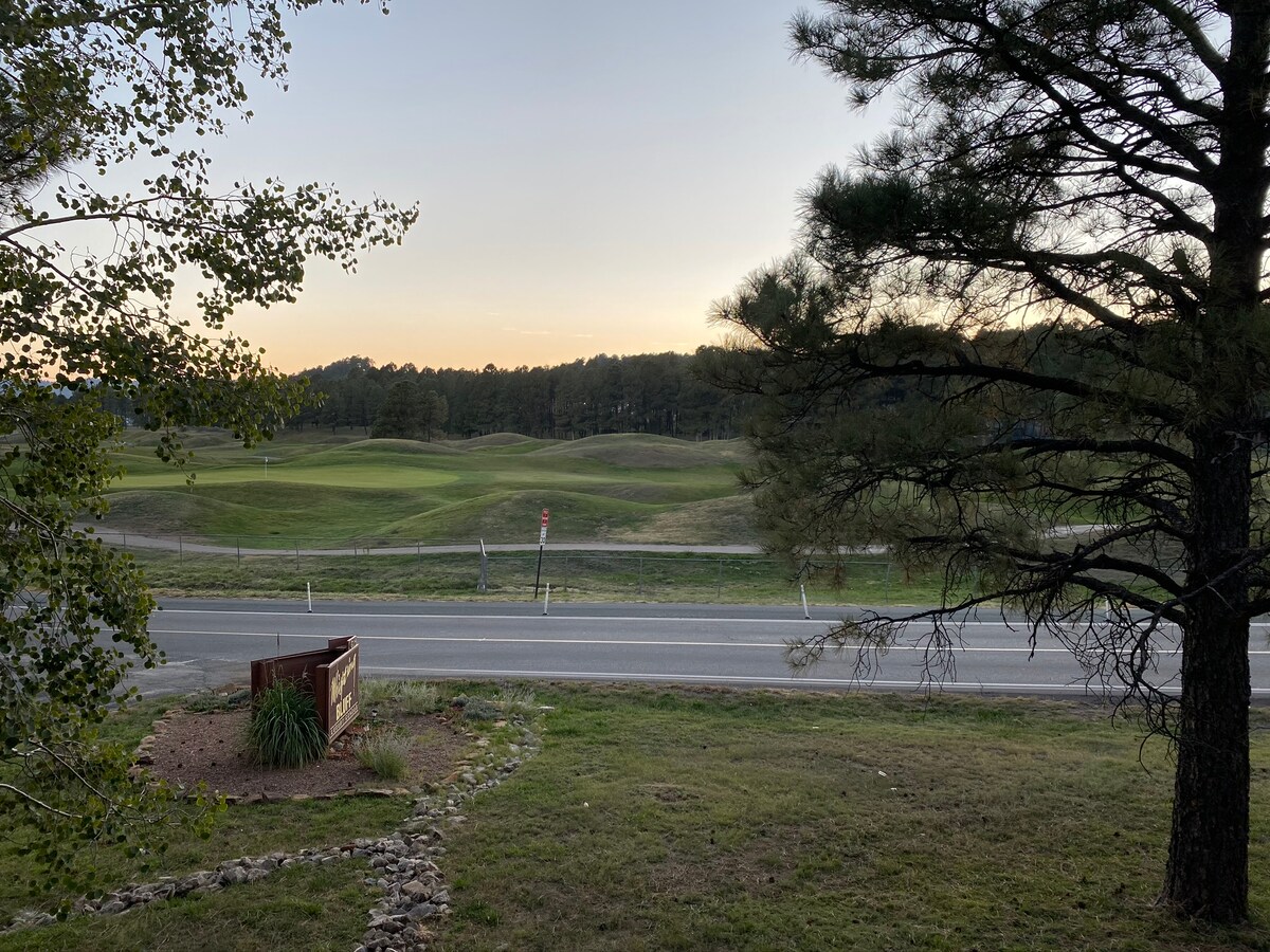 A scenic view captures lush green hills of a golf course at dusk, framed by tall trees. The foreground shows a neatly maintained sign and a grassy area leading to the road, creating a peaceful ambiance.