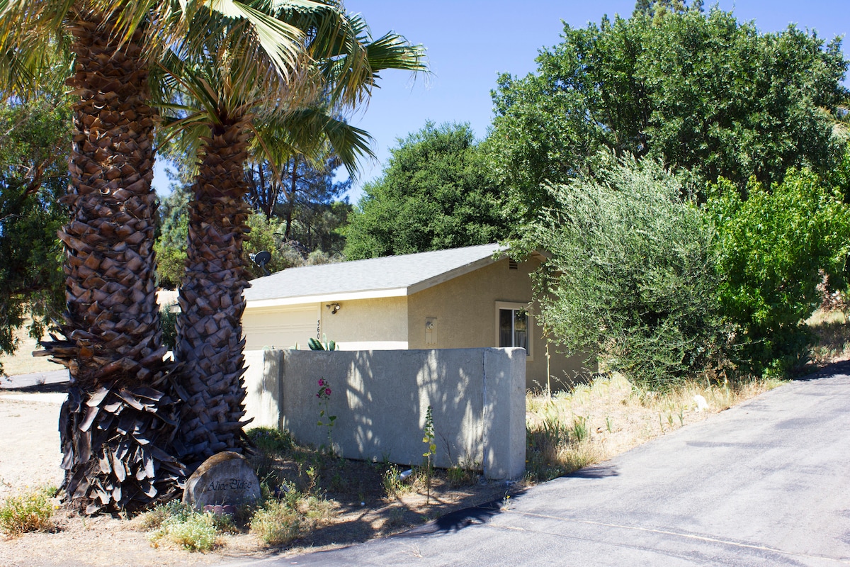 The exterior of the guesthouse is depicted, featuring a low stone wall and mature palm trees framing the entrance. Lush greenery surrounds the structure, blending with the natural landscape. A paved road is visible, leading towards the inviting entrance of the guesthouse.
