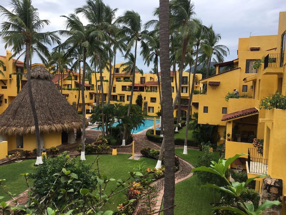A vibrant condominium complex is surrounded by lush greenery and palm trees. The yellow buildings create a warm atmosphere, with a thatched-roof palapa visible in the center. A swimming pool is situated among landscaped gardens, connecting walking paths through the area.