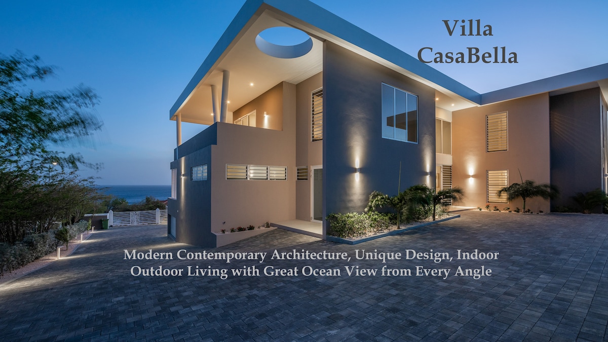 The exterior of Villa CasaBella is showcased, highlighting modern architectural design with clean lines and large windows. The illuminated pathway guides the eye towards the entrance, while the ocean view is visible in the background, emphasizing the villa's connection to its natural surroundings.