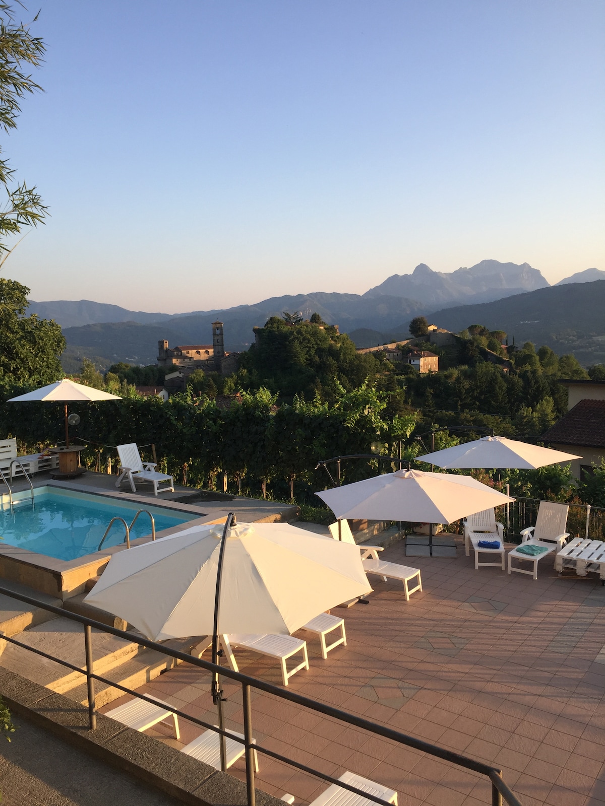 An outdoor area features a pool surrounded by sun loungers and umbrellas. The backdrop includes lush greenery and a scenic view of mountains and the village. The late afternoon light creates a calming atmosphere, enhancing the enjoyment of the space.