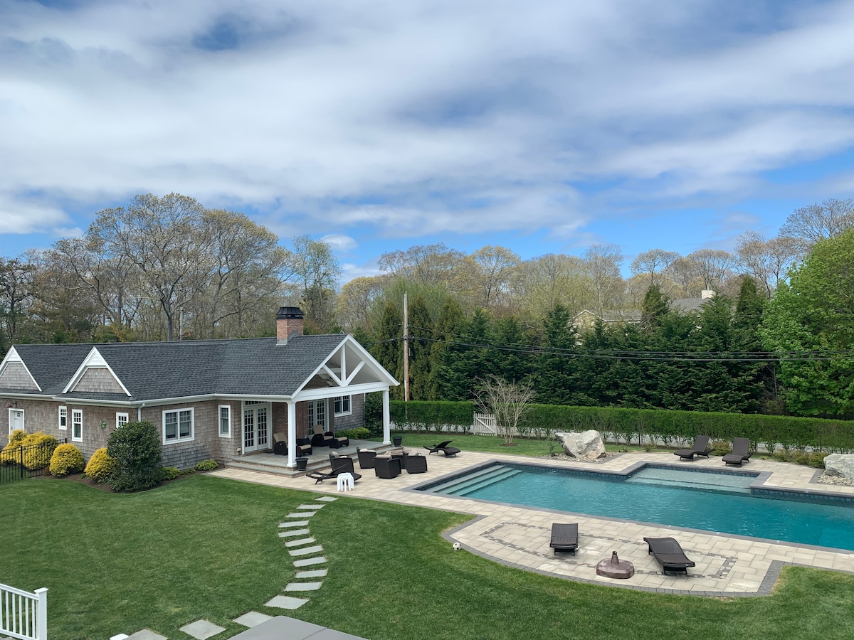 The image captures a spacious outdoor area featuring a large, heated swimming pool surrounded by lounge chairs. A well-maintained lawn is visible, along with a charming cottage that includes an inviting porch. Lush greenery and trees form a serene backdrop.