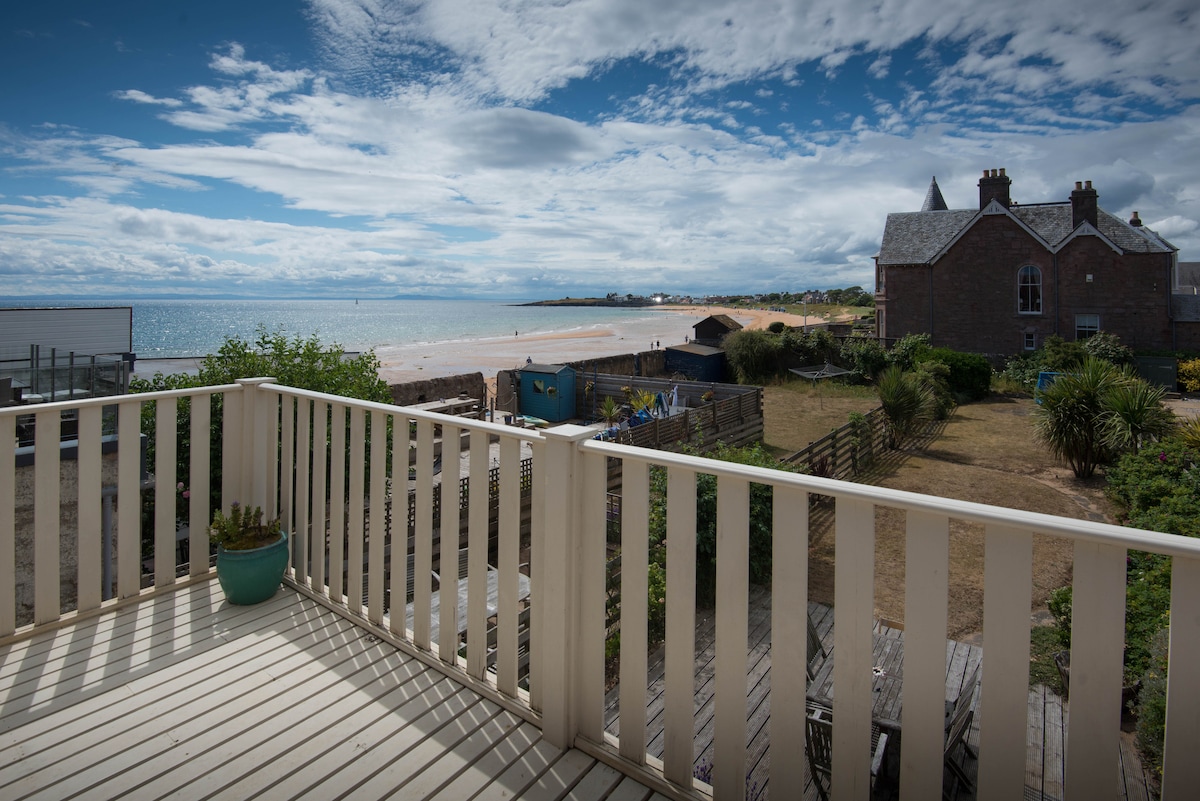 A balcony offers expansive views of the beach and sea, with sandy shores stretching into the distance. Surrounding gardens and buildings are visible, framed by a clear blue sky dotted with soft white clouds.