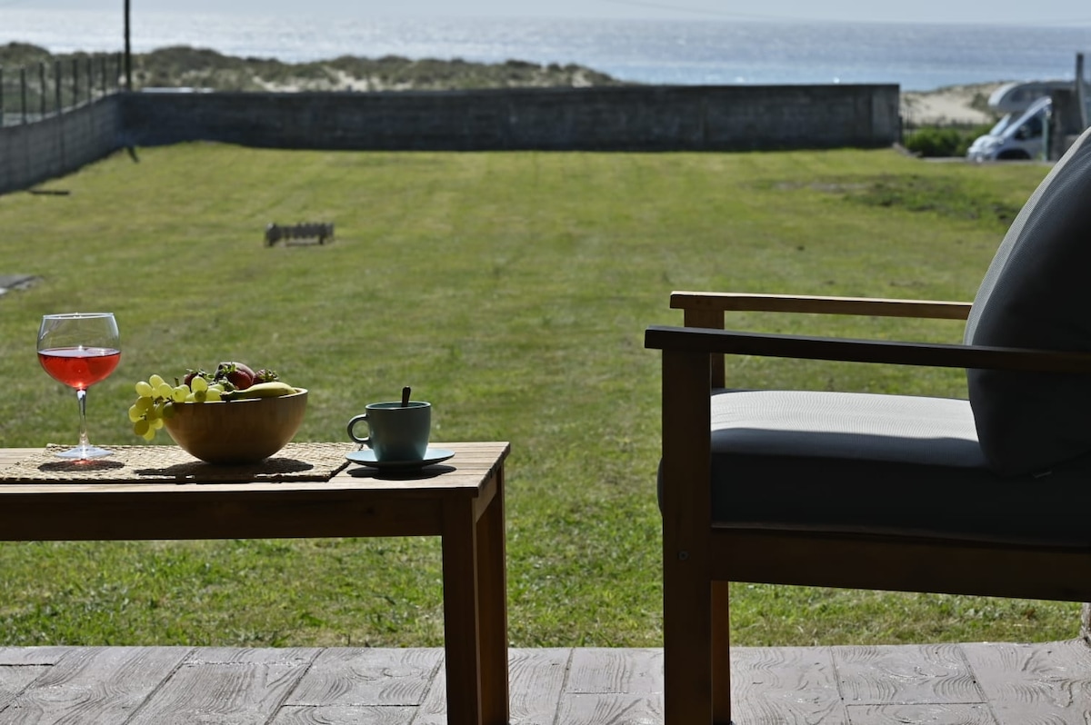 A wooden table with a glass of beverage and a bowl of fruit sits beside a comfortable chair. The view extends to a grassy area leading towards the beach, with the ocean visible in the background under a clear sky.