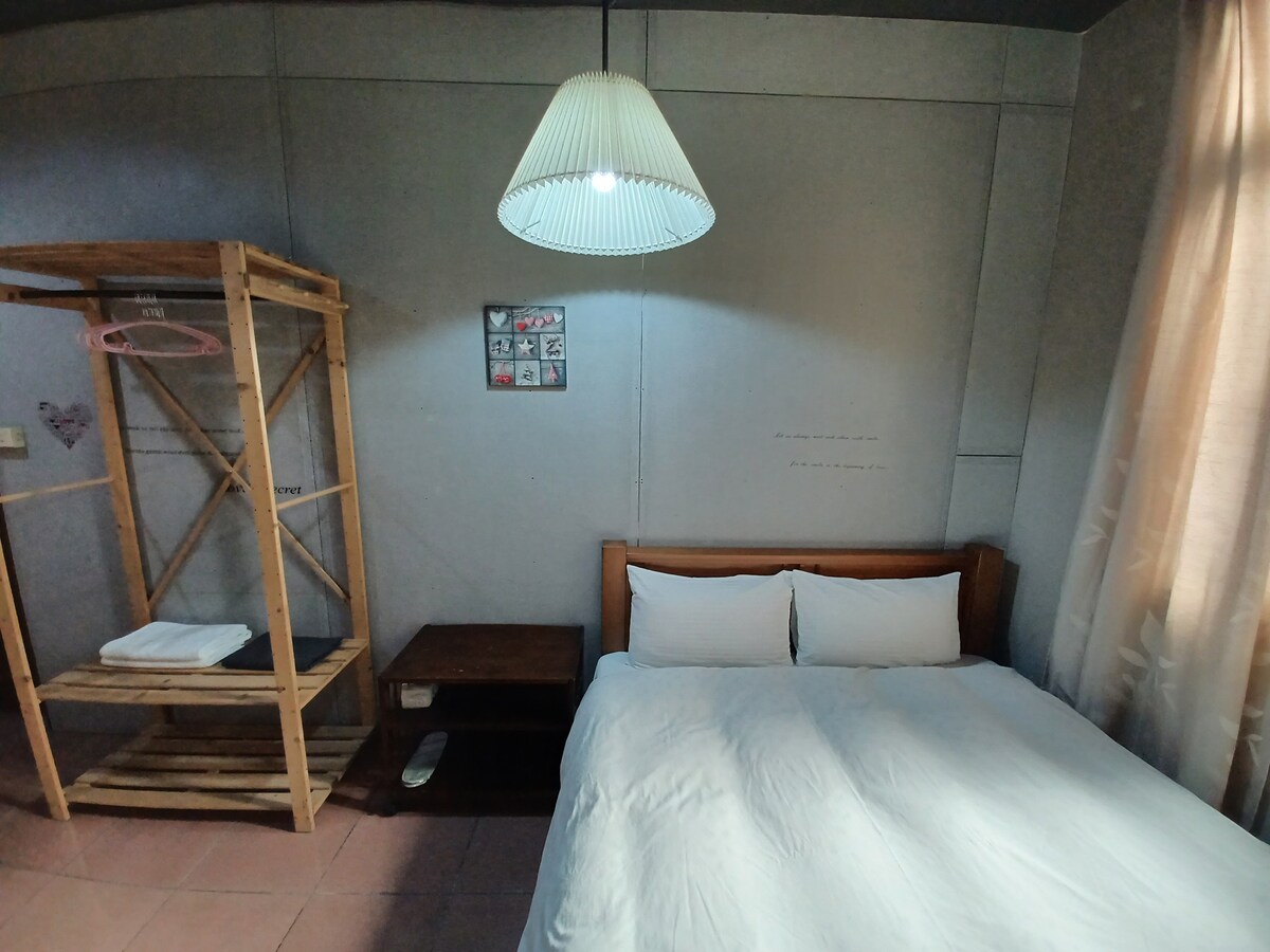 A simple and clean room is featured, with a spacious bed dressed in white linens. A wooden side table is positioned next to the bed, and a bamboo shelf holds neatly folded towels. Soft lighting from a pendant lamp illuminates the space.