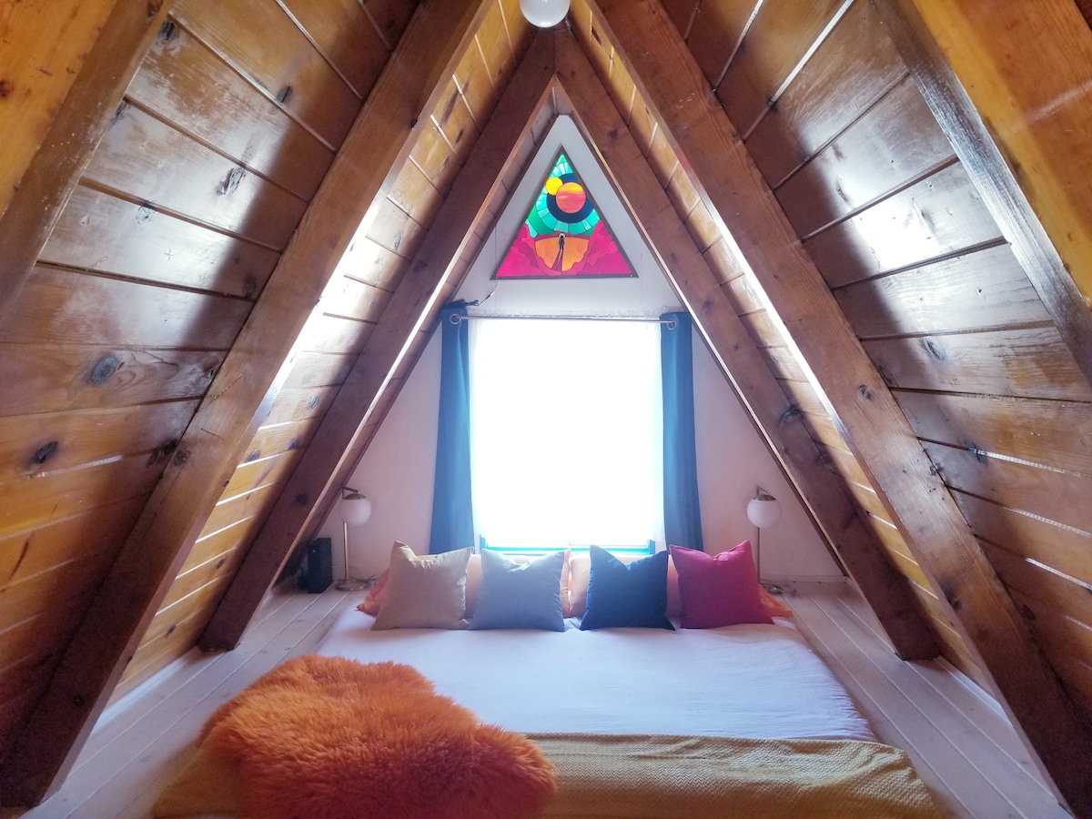 Upstairs loft with sunken king bed (yes, full depth mattress!), duvet and lots of fluffy pillows. View of the mountains from the window. Sci fi stained glass lamp made by your host above. Integrated storage and power/usb in pine bed frame.