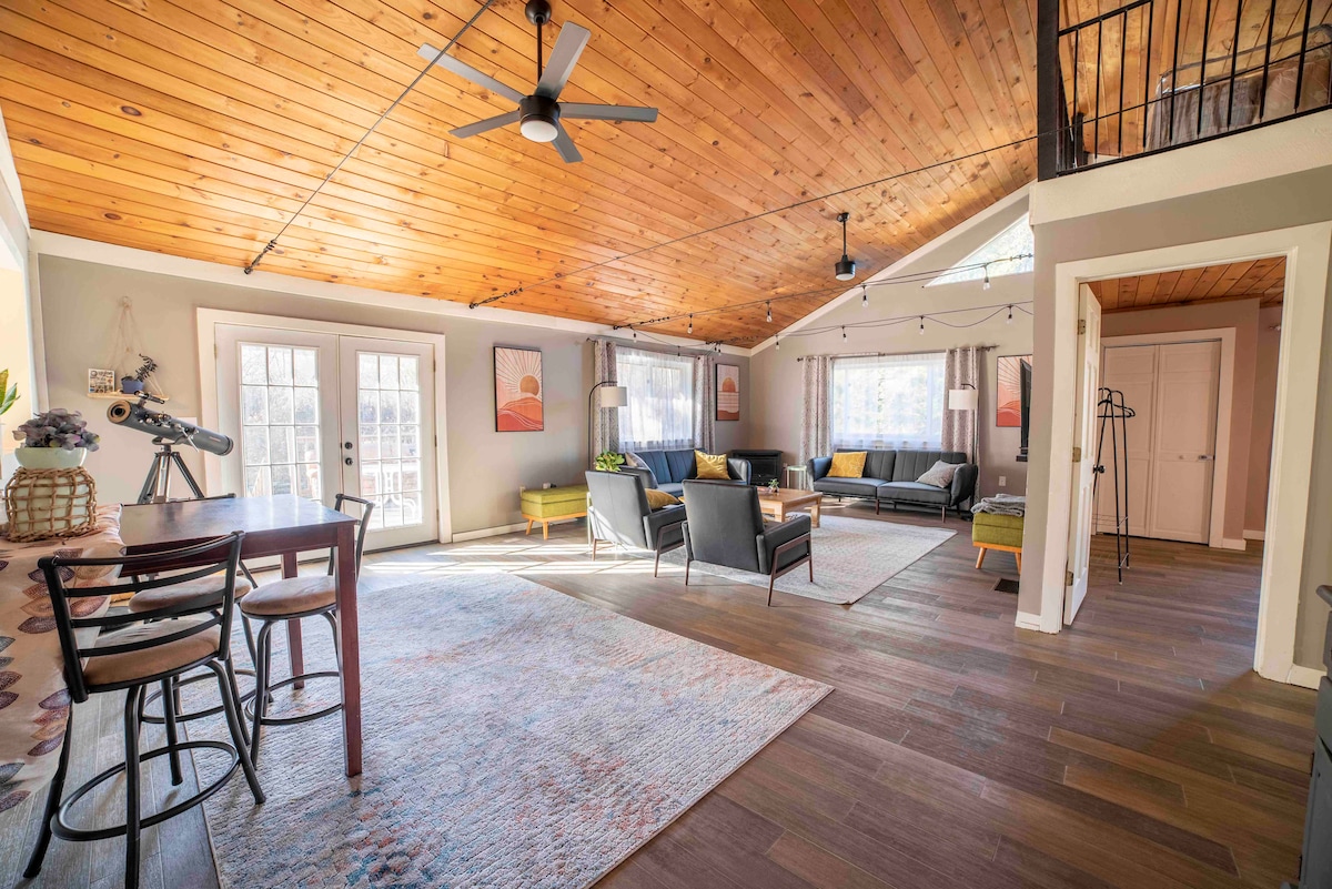 Bright living space features high, wooden ceilings and large windows allowing natural light. Comfortable seating arrangements are provided by several couches and chairs, complemented by an area rug. Access to a deck is offered through two glass doors, enhancing the spacious feel of the room.