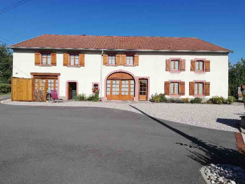 A renovated farmhouse is displayed with a smooth gravel driveway leading to a light-colored facade. Wooden shutters frame the windows, and a large archway entrance is visible. Surrounding greenery includes low shrubs and a patio area is indicated on the left.