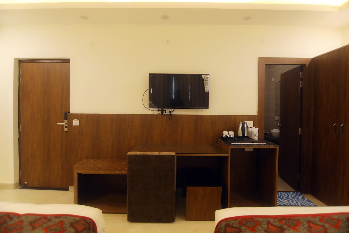 A functional room setting is presented with a wall-mounted television and a compact work desk. A coffee maker and accessories are neatly arranged on the counter. The doors lead to a bathroom and a hallway, while the light-colored walls create a spacious feel.