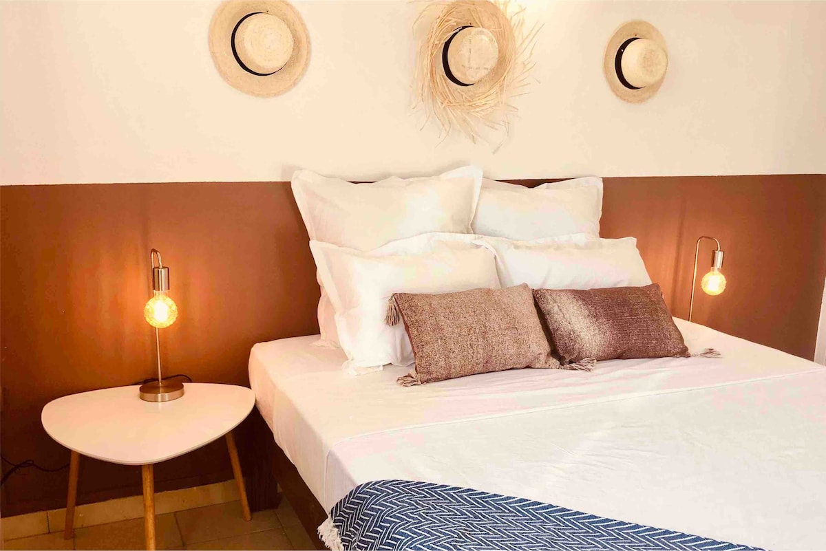 A well-arranged sleeping area is seen, featuring a spacious bed adorned with fluffy white pillows and a textured throw blanket. Soft golden lighting is provided by two bedside lamps, while decorative hats hang on the wall above the headboard, contributing to a relaxed ambiance.