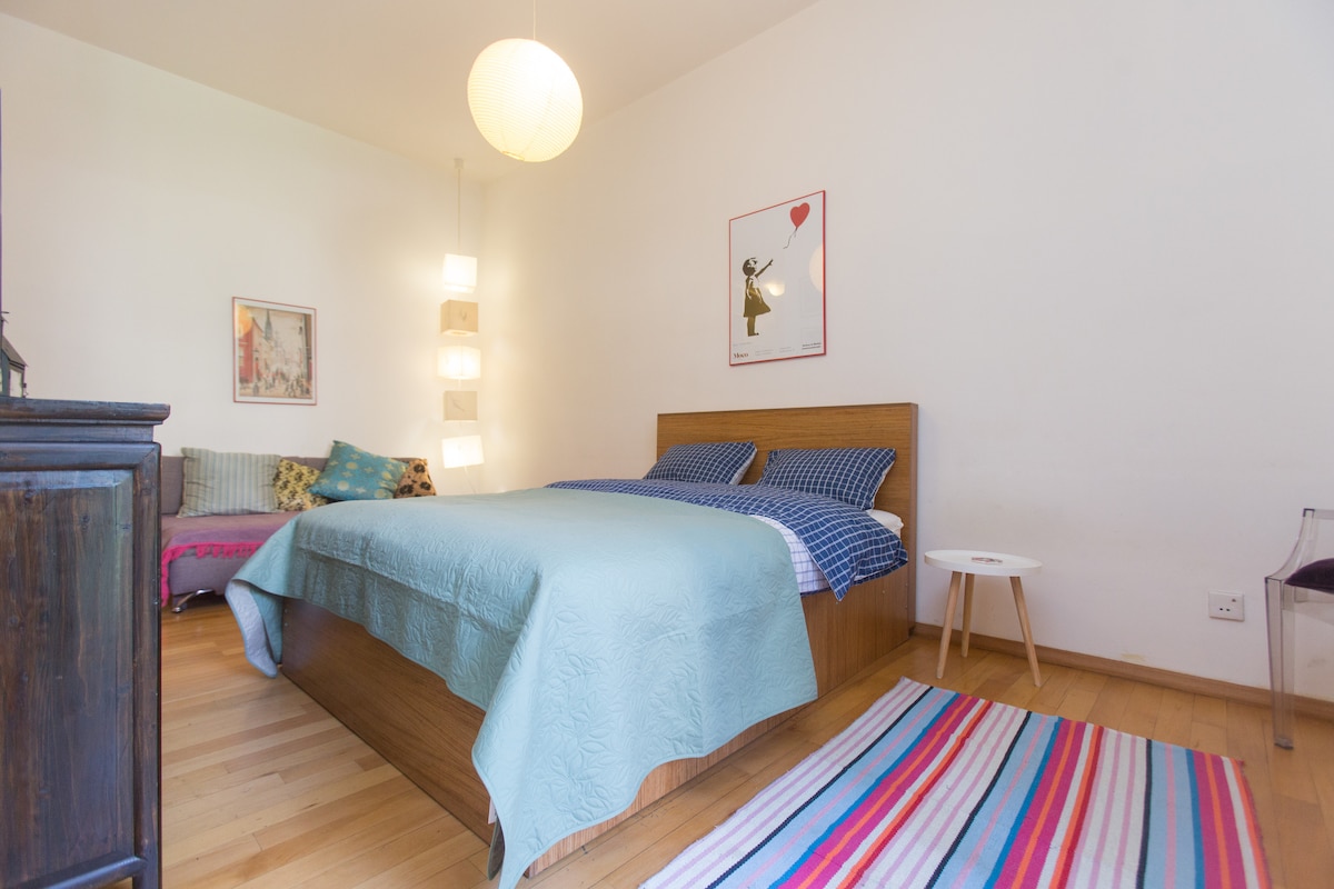 A spacious sleeping area is provided with a wooden bed dressed in a light blue coverlet. Decorative lighting hangs from the ceiling, and a colorful striped rug adds warmth to the hardwood floor. Artwork is displayed on the walls, contributing to the inviting environment.