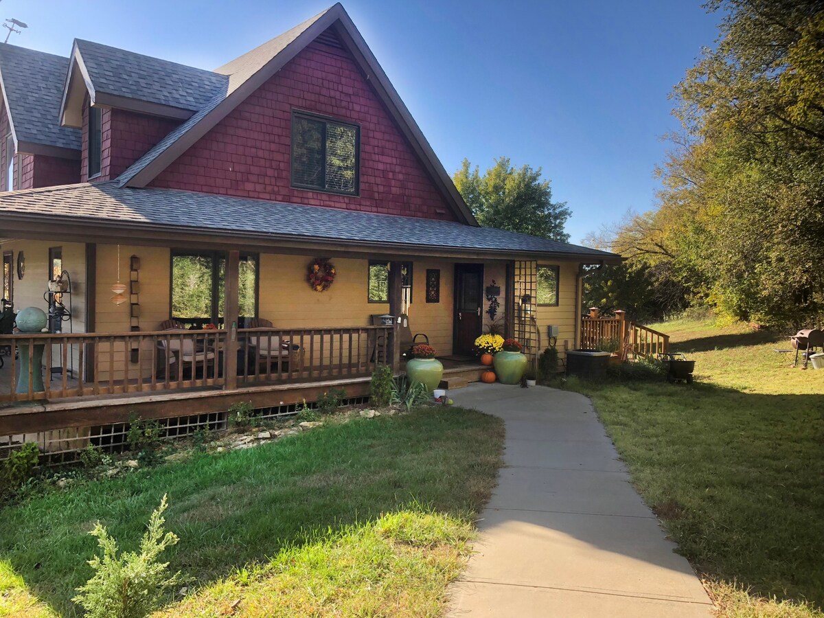 A welcoming entrance is highlighted by a spacious porch adorned with seasonal decorations. Lush greenery surrounds the pathway, and outdoor seating provides inviting spots to relax. The charming house features a combination of brick and wooden siding, with large windows opening to the surrounding landscape.