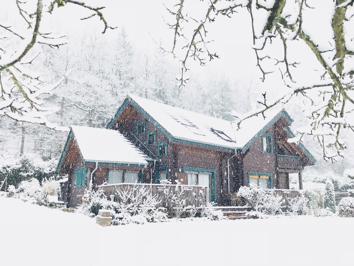 A charming chalet is set against a snowy backdrop, showcasing a wooden exterior with teal accents. The property features a spacious deck partially obscured by fresh snow, surrounded by white-draped trees and scenery, creating a serene winter atmosphere.