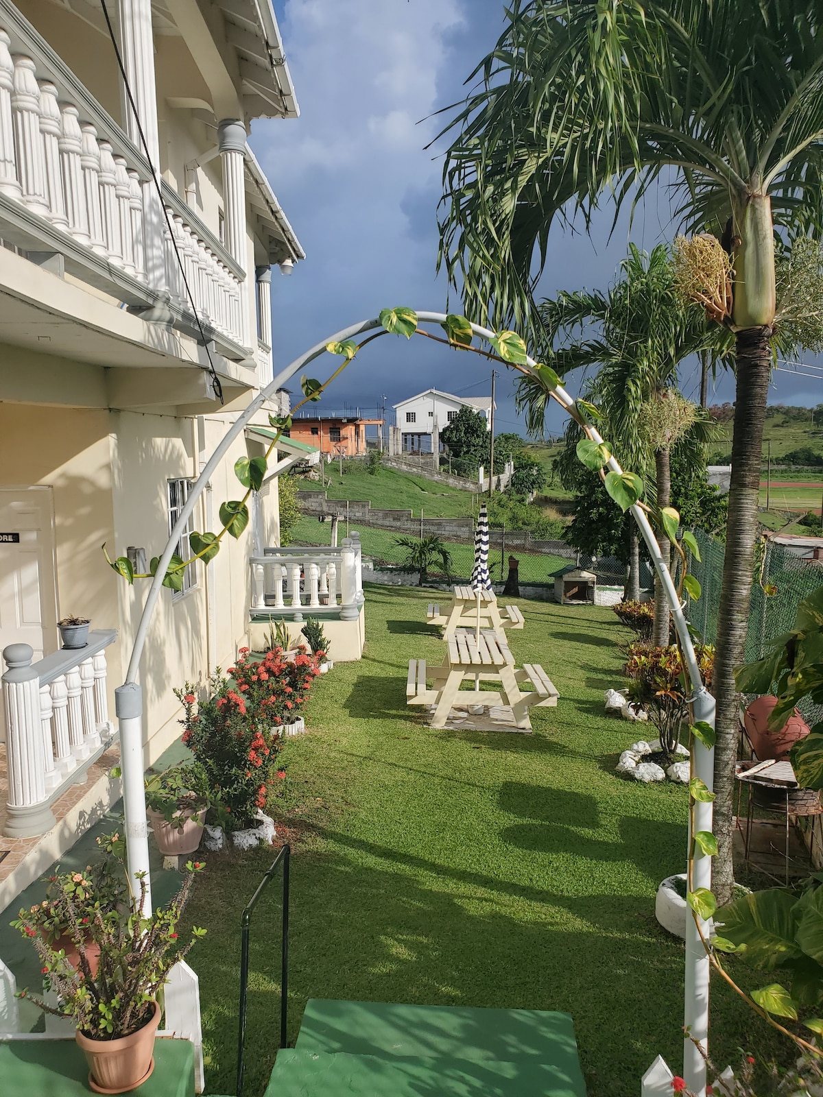 A well-maintained garden area includes a neatly trimmed lawn with palm trees and flowering plants. A white archway adorned with green vines leads to a picnic-style wooden table and benches set in the distance. The sky is partly cloudy, completing the serene outdoor view.