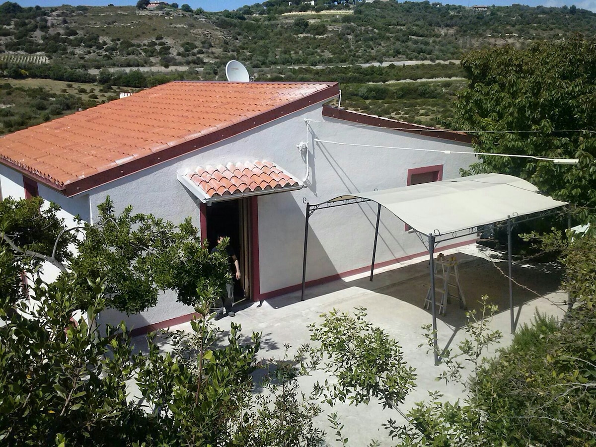 A single-story villa is showcased featuring a terracotta roof and outdoor shaded area. Surrounding foliage enhances the natural setting, while the entrance is framed by an inviting pathway. The landscape includes gentle hills, providing a serene backdrop.