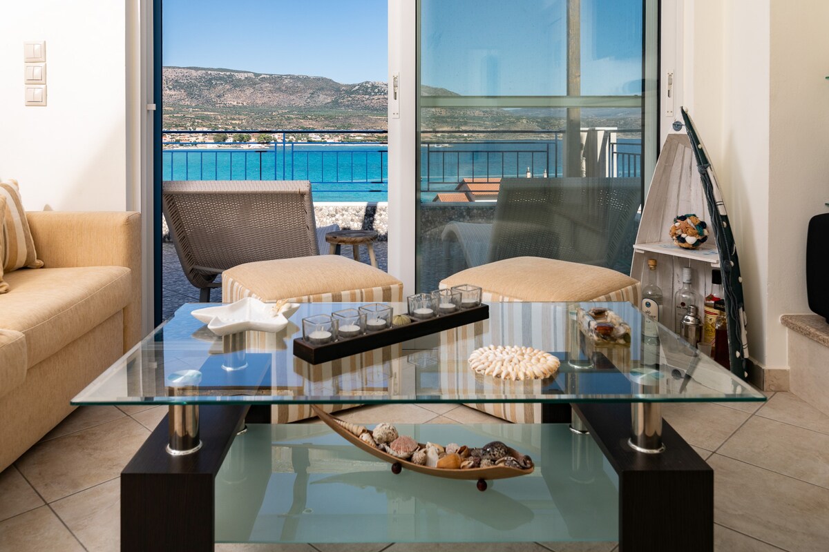 A modern living area features large glass doors that invite natural light and offer views of the sea. A glass coffee table showcases decorative shells and candles, while comfortable seating options are arranged for relaxation. A balcony is visible, enhancing the coastal ambiance.