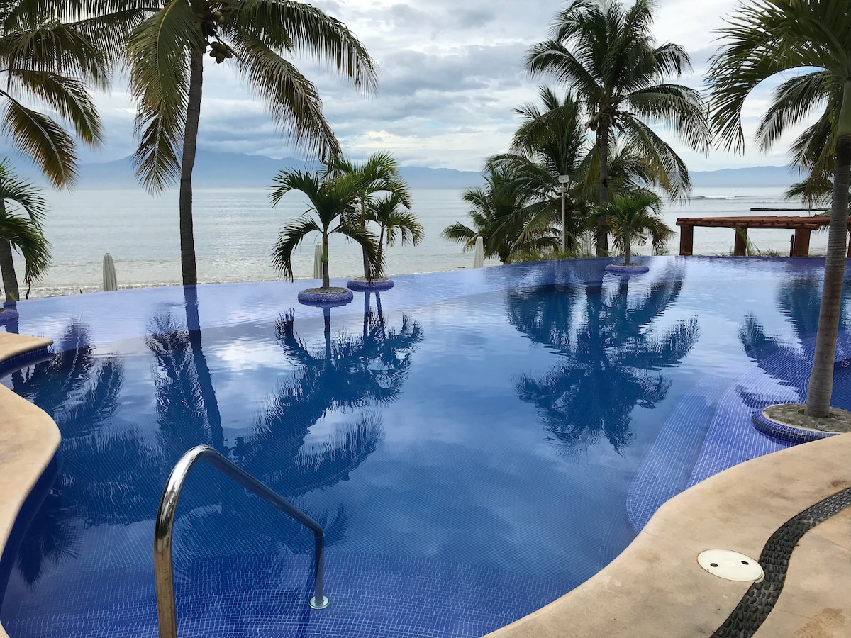 An infinity pool is seen seamlessly merging with the ocean in the background, surrounded by lush palm trees. The water's surface reflects the tranquil sky, creating a serene environment that invites relaxation.