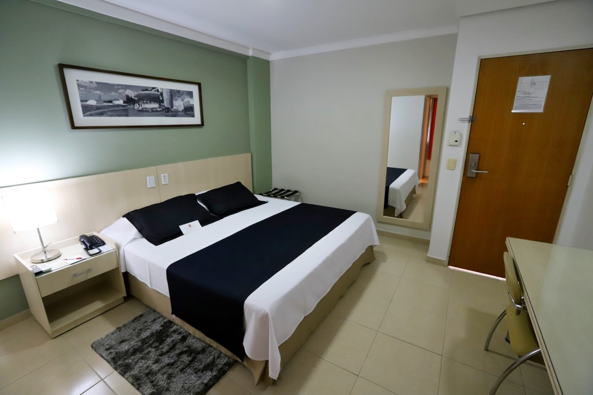 A well-arranged room features a spacious bed with dark linens and light-colored bedding. A bedside table is placed beside the bed, and a mirror reflects the light from a lamp. A door and a desk complete the simple yet functional layout of the space.