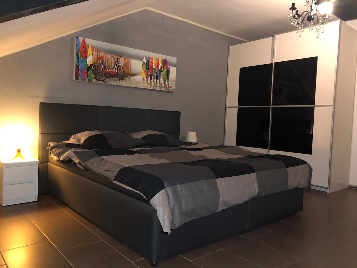 A spacious bedroom features a large box spring bed adorned with a patterned duvet. A stylish piece of artwork is displayed above the bed, complemented by a bedside table with a lamp. Mirrored wardrobe doors reflect light, enhancing the room's sense of space.