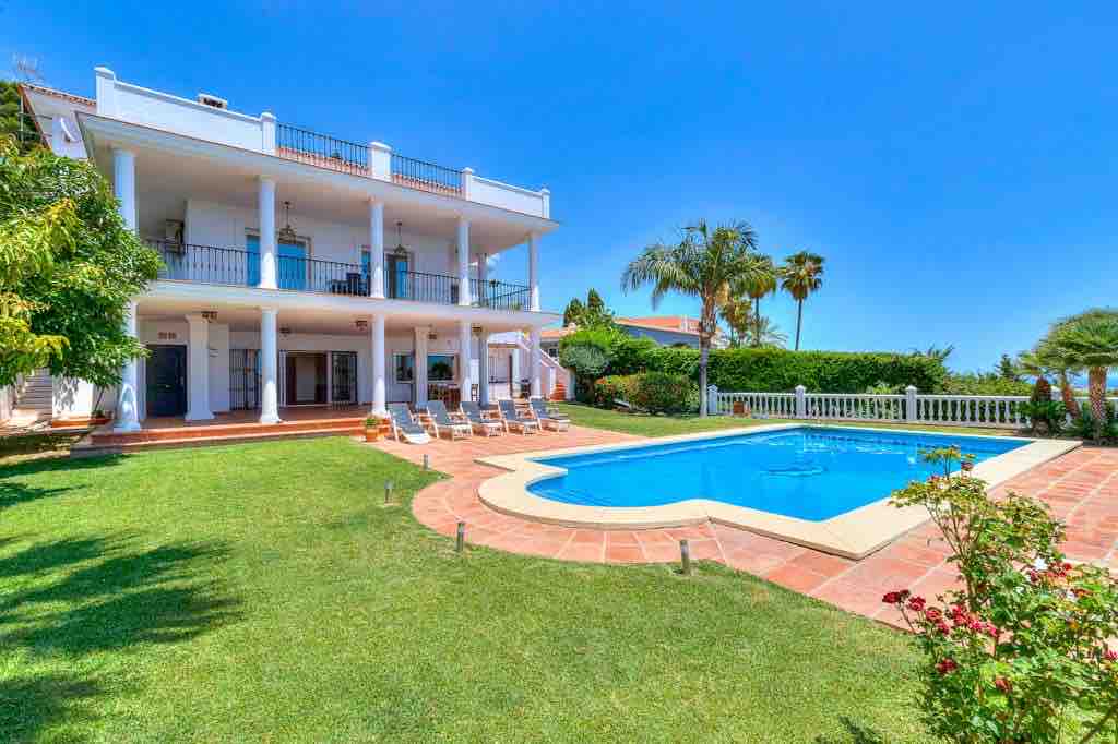 A spacious villa is depicted with multiple balconies overlooking a well-maintained lawn and a private pool. Lounge chairs are arranged around the pool, surrounded by palm trees and colorful garden plants. Clear blue skies provide a bright backdrop to the scene.