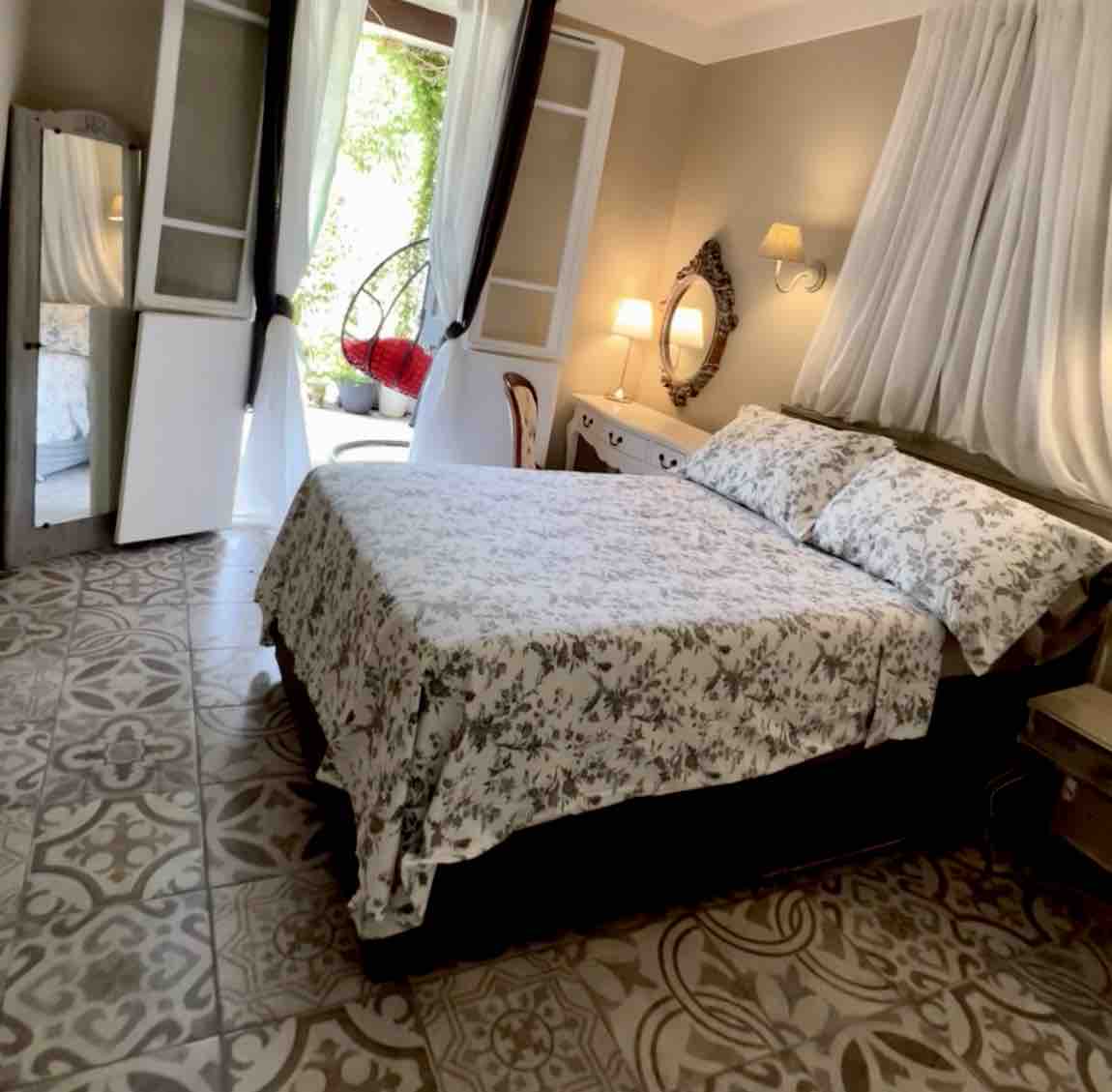A spacious bedroom is furnished with a comfortable bed dressed in a floral pattern. Natural light filters through sheer curtains, illuminating the patterned floor tiles. A cozy seating area outside the window invites relaxation, and a full-length mirror is positioned nearby.