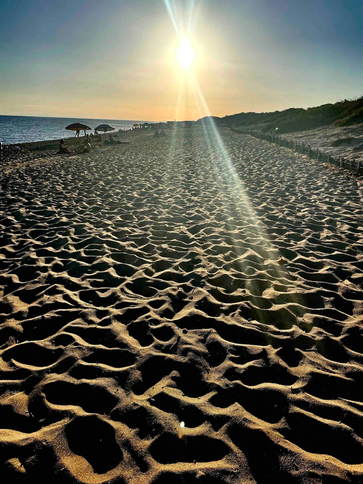 A sandy beach stretches along the shoreline, illuminated by sunlight. The golden sand reveals captivating patterns from the ebb and flow of the tide, while distant silhouettes of umbrellas provide shade. Sparse vegetation outlines the beach, framing a serene coastal scene.