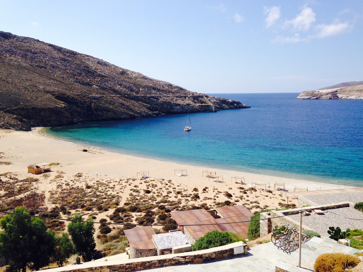 A sandy beach stretches along the shoreline, meeting azure waters under a clear blue sky. Gentle hills rise in the background, providing a rugged contrast to the beach scene. A sailboat floats quietly in the distance, adding to the serene atmosphere.