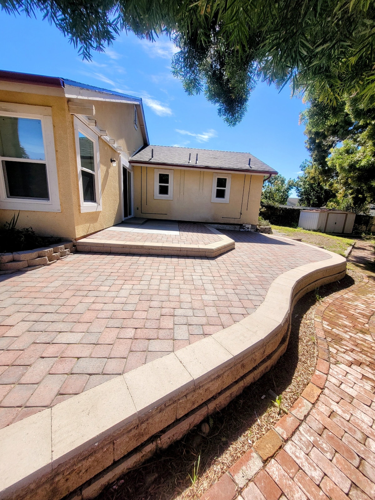 A paved patio area is bordered by smooth stone, providing ample space for outdoor seating or activities. Sunlight filters through nearby trees, casting gentle shadows. The adjacent building is visible, enhancing the spaciousness of the area.