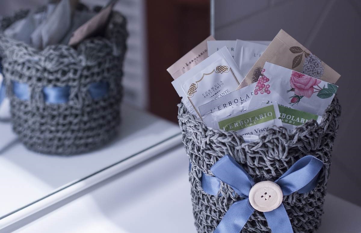 A woven gray basket is displayed in front of a mirror, featuring various individually packaged herbal products. A blue ribbon and button are tied around the basket, adding a touch of detail to its design.