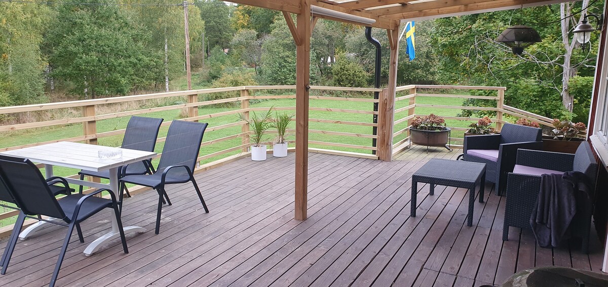 A spacious wooden deck is shown, featuring a table with four chairs and comfortable seating arrangements. Lush greenery surrounds the area, providing a peaceful outdoor space. Solar lights are visible in the corners, offering subtle illumination.