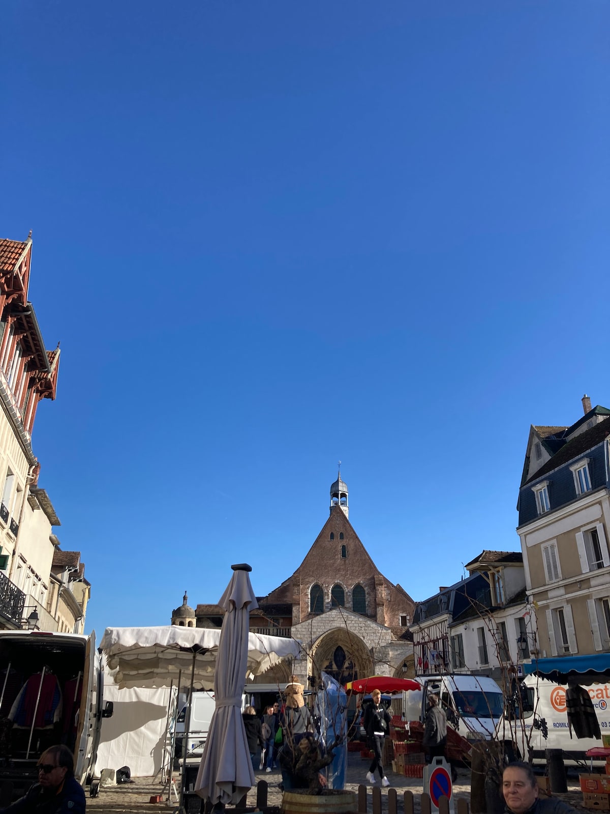 A clear blue sky fills the frame above a bustling market scene. The architectural details of a notable building can be seen in the background, showcasing a distinctive roofline. Stalls and people are visible, contributing to the vibrant atmosphere of the market.