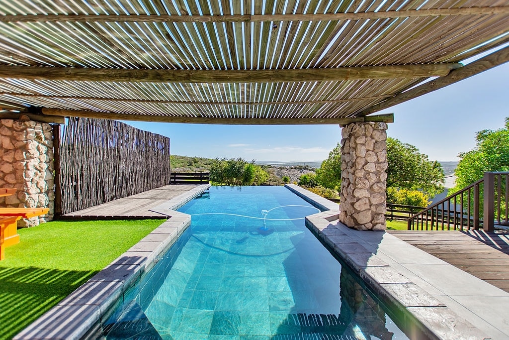 A serene heated pool is framed by natural stone and wooden structures, creating a shaded seating area. The pool stretches into the distance, with views of lush greenery and a clear sky visible beyond. The reflection of the surrounding nature can be seen on the water's surface.