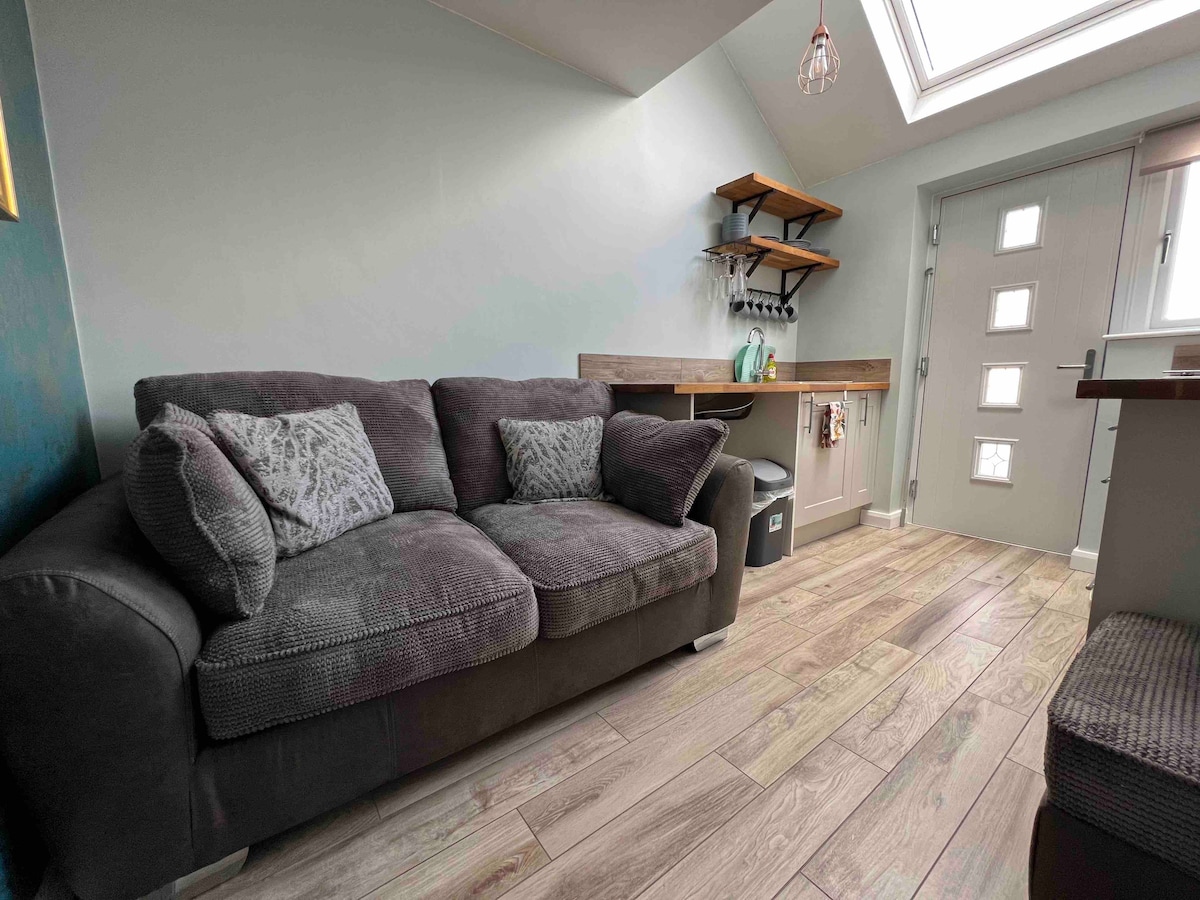 A comfortable sofa with decorative cushions is positioned against the light-colored wall. A wooden shelf rests above a countertop area, complementing the tiled floor. Natural light pours in through a skylight, illuminating the inviting living space near the entrance.