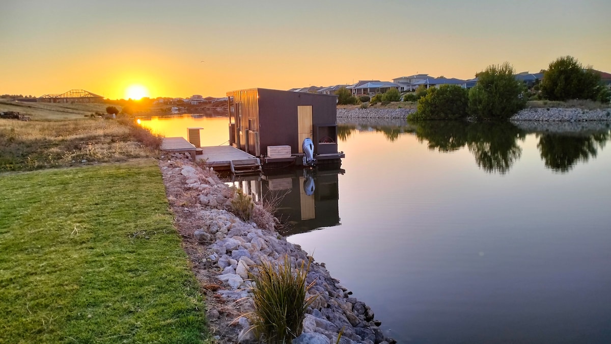 A tranquil riverside scene captures a modern structure on a jetty, reflecting in calm waters at sunset. Lush green grass leads towards the water, framed by rocky landscaping. Soft hues of orange and yellow from the setting sun create a serene backdrop against the still water.