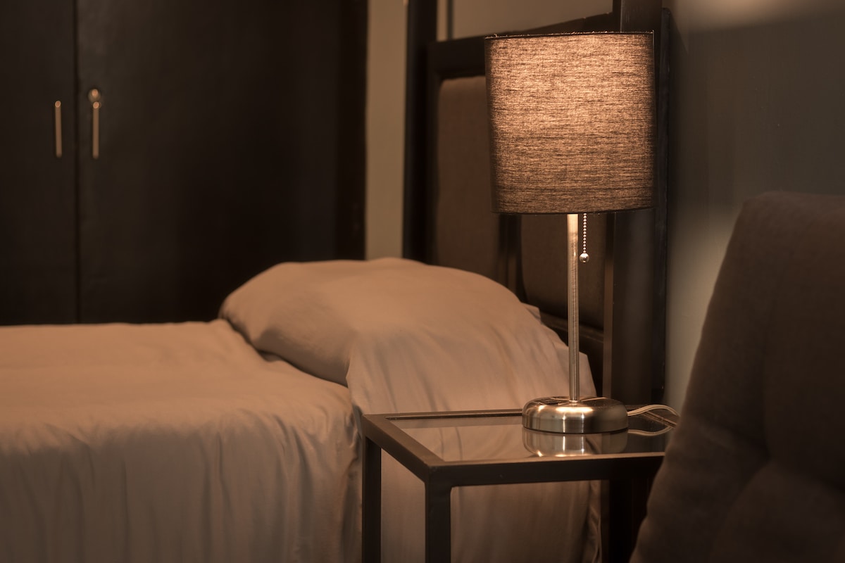 A cozy sleeping space features a spacious bed dressed in neutral linens. A simple bedside table holds a stylish lamp that gently illuminates the area. The dark walls provide a contrasting backdrop, enhancing the room's calming atmosphere.