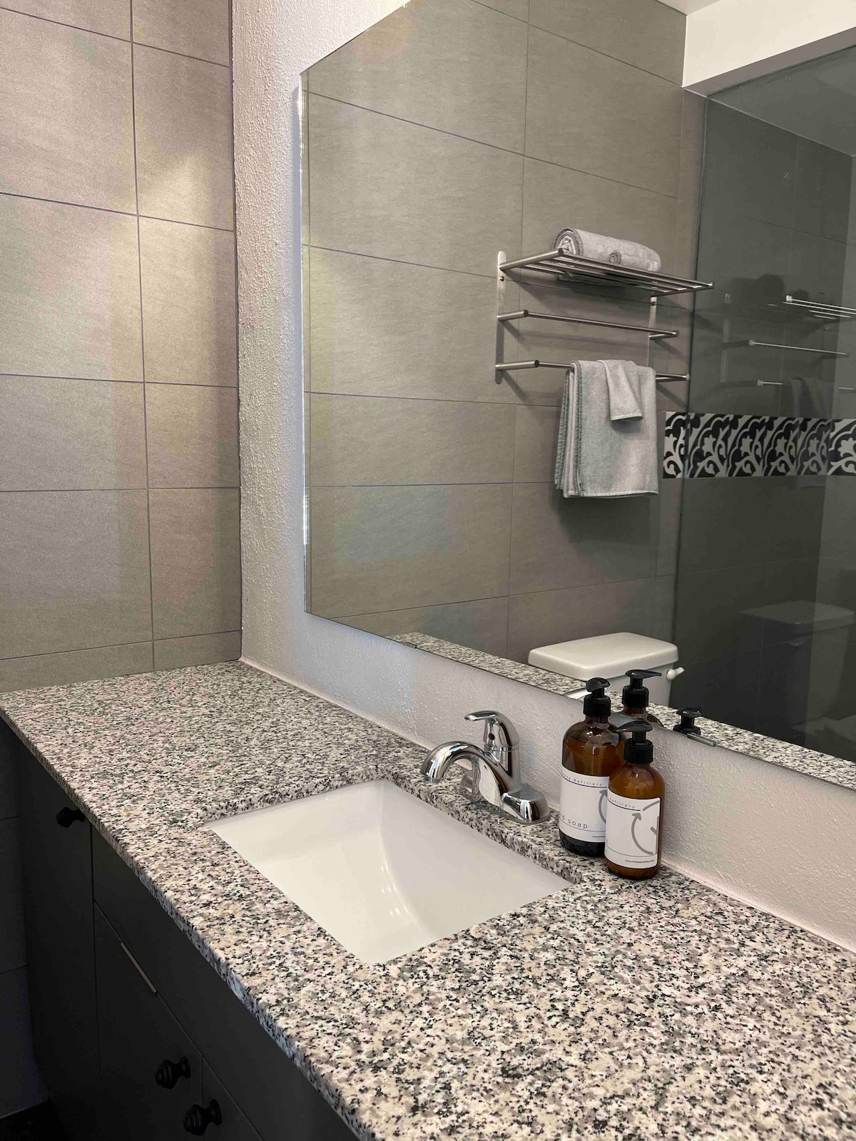 The bathroom features a modern design with tiled walls and a granite countertop. A single sink is positioned beneath a large mirror. Shelving holds neatly arranged towels, and a set of soap bottles is placed on the countertop, highlighting the space's functionality.