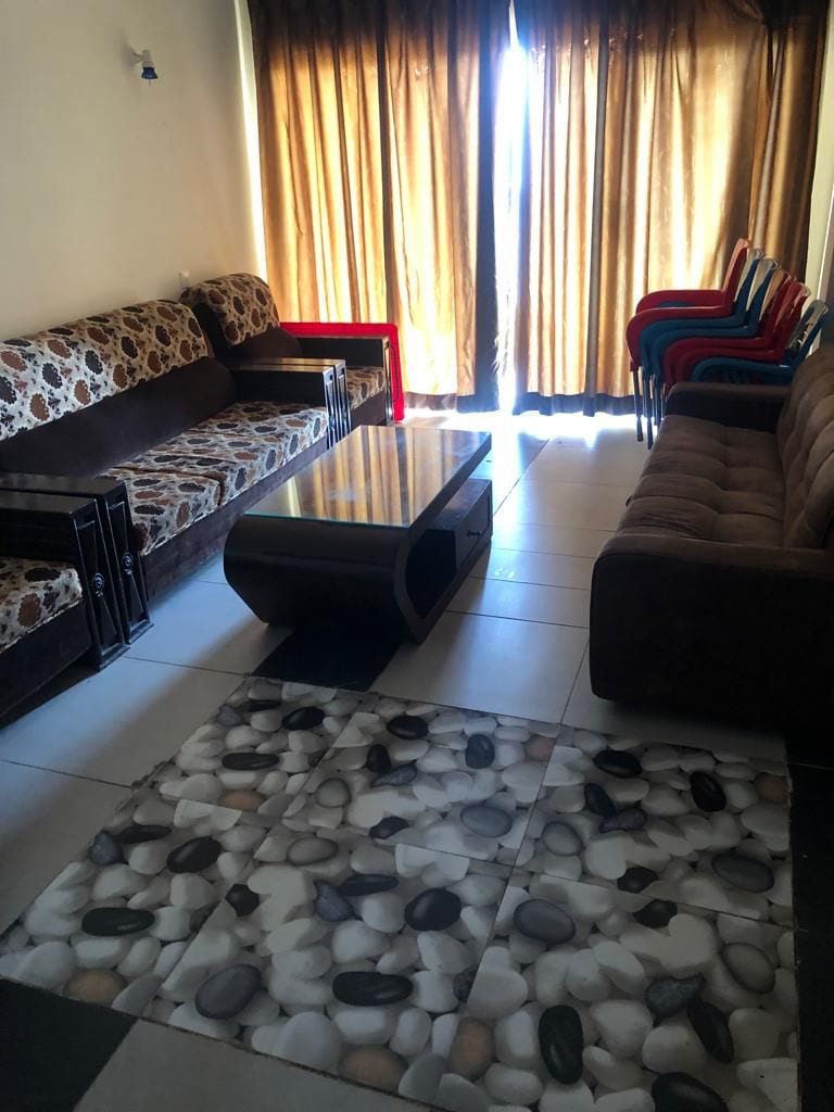 A cozy TV lounge area is furnished with patterned sofas and a brown couch, arranged around a low coffee table. Light filters through the floor-to-ceiling curtains, highlighting a unique stone-patterned floor that adds texture to the space.